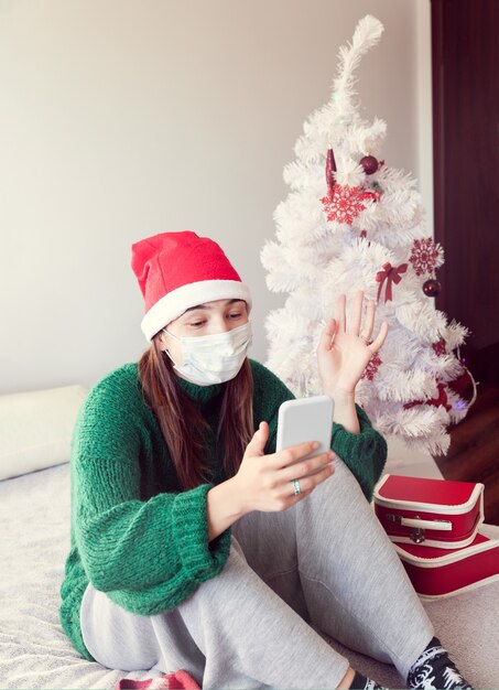 Lockdown for Christmas with girl onlane. Girl with a face mask and in a hat of Santa Claus is chatting by phone