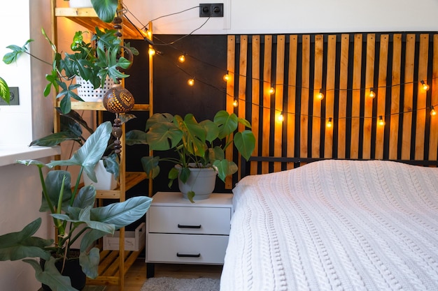 Спальня в стиле лофт интерьер черная стена с деревянными решетками металлическая кровать ретро лампочки гирлянда горшки растения на трапецеобразном окне на чердаке современный зеленый дом