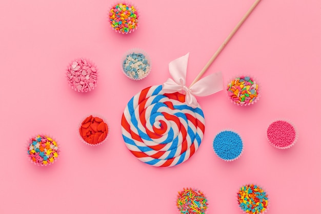 Lollipop and sugar sprinkles in paper bowls on pink background, top view