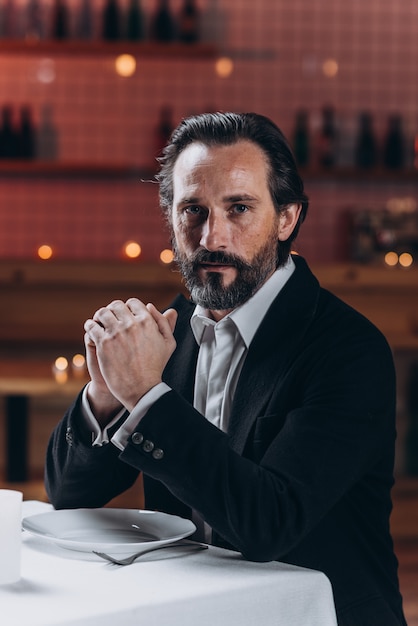 Lonely bearded man in a suit sits alone at a table in a restaurant and looks into the camera.