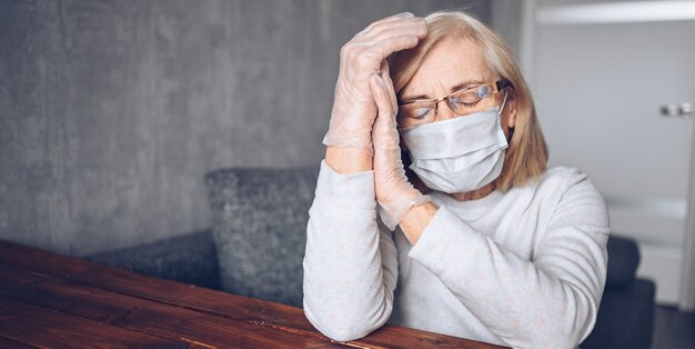 Lonely depressed sad older elderly senior woman in face protective mask, glasses, and medical gloves sitting at home self isolation quarantine during coronavirus COVID19 pandemic. Stay home concept