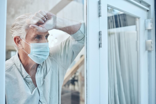 Lonely face mask and senior man in the house for quarantine protection and isolation during pandemic Depression frustrated and sad elderly guy with covid looking outside the window at his home