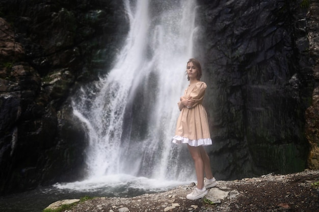 Lonely girl in a short beige dress stands next to the waterfall