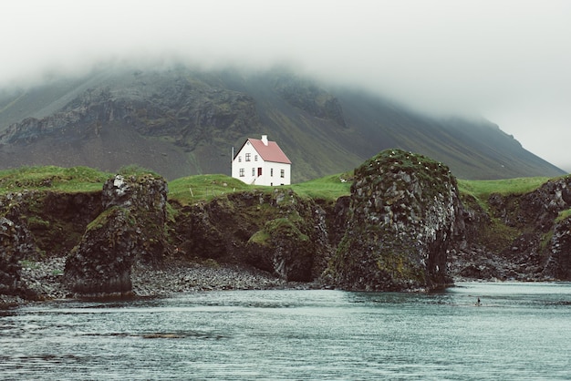 Одинокий исландский дом с красной крышей на морском побережье с зеленой травой, луговыми скалами и туманным небом Природный ландшафт путешествия по Исландии