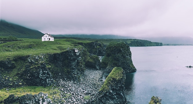 Lonely icelandic house with red roof on the sea coast with green grass meadow rocks anf foggy sky Natural Iceland travel landscape