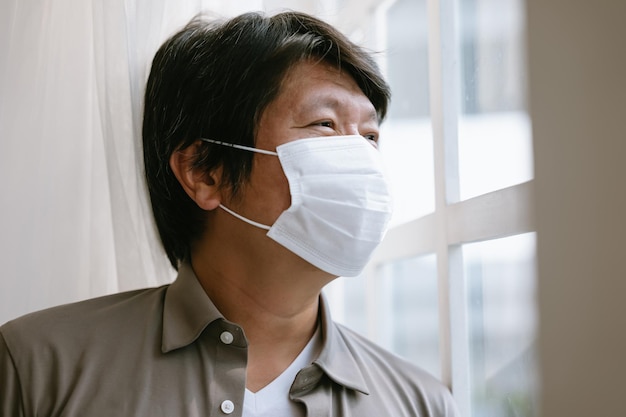自己検疫のために自宅の窓から見ている医療用フェイシャルマスクを着た孤独な韓国人男性フェイスマスクを着た不幸な男性はコロナウイルスに苦しんでいます検疫コロナパンデミックの概念