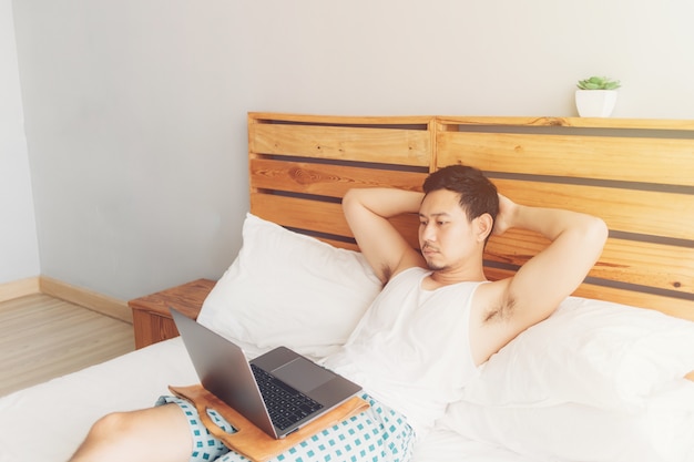 외로운 남자는 그의 아늑한 침대에서 자신의 노트북을 사용하고 있습니다. 프리랜서 작업 라이프 스타일의 개념입니다.