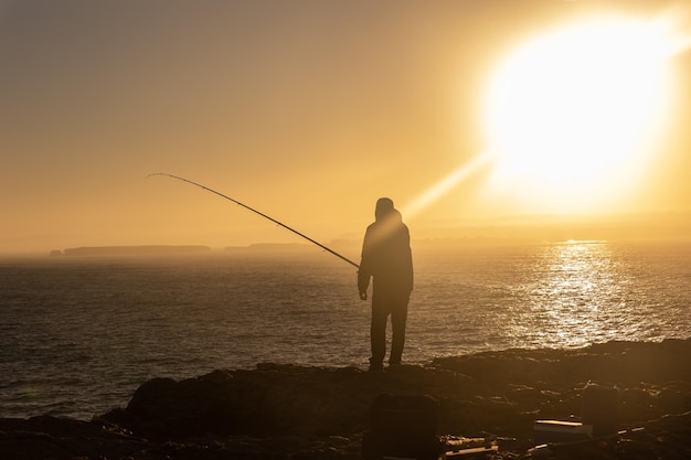 Одинокий мужчина стоит на вершине скалы и ловит рыбу в море на закате