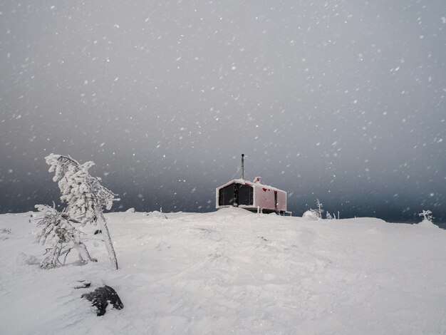 夜の劇的な空の下で冬の雪に覆われた斜面にある孤独な赤いゲストハウス 山の上の観光客の家のための冬の避難所