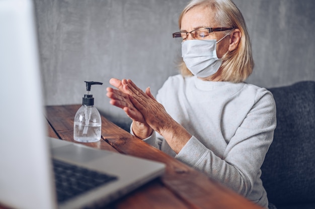 Lonely sad older senior woman in face medical mask using hand antibacterial liquid sanitizer with laptop computer at home self isolation quarantine during coronavirus COVID19 pandemic. Stay home