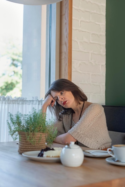 Lonely sad pensive or tired woman sits at table in cafe with cup of coffee and dessert Young woman alone in cozy cafe