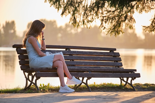 Lonely sad woman sitting alone on lake shore bench on warm summer evening. Solitude and relaxing in nature concept.