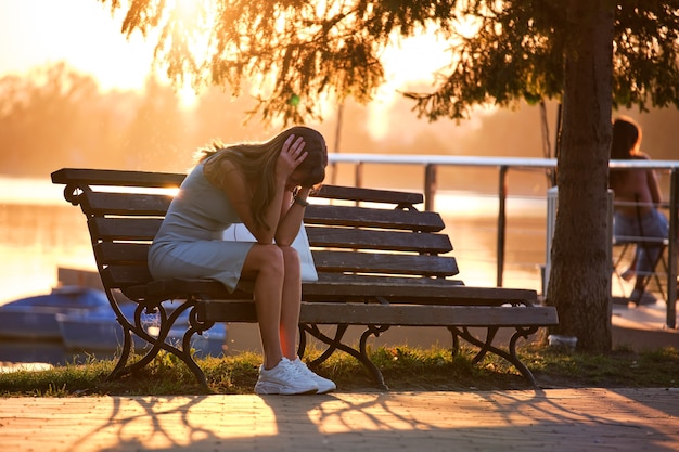 Lonely unhappy young woman sitting alone on park bench on warm summer evening. Solitude and relaxing in nature concept.