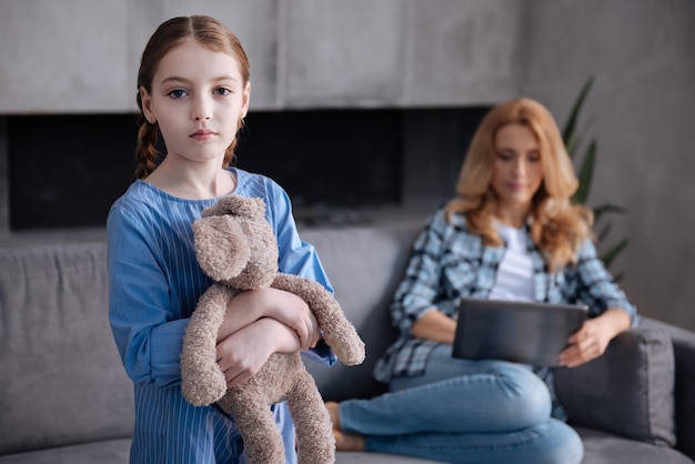 Lonely upset melancholy kid waiting for parent care at home and hugging fluffy toy while mother working with tablet