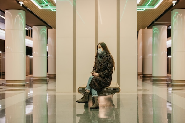 A lonely woman in a medical face mask to avoid the spread of coronavirus is sitting with a smartphone on the subway platform. A girl in a surgical mask is keeping social distance in the metro.