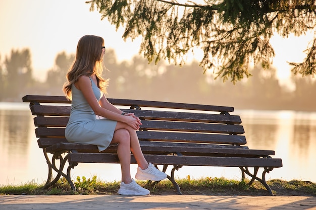Одинокая женщина, сидящая одна на скамейке на берегу озера в теплый летний вечер. Уединение и расслабляющий отдых на природе.
