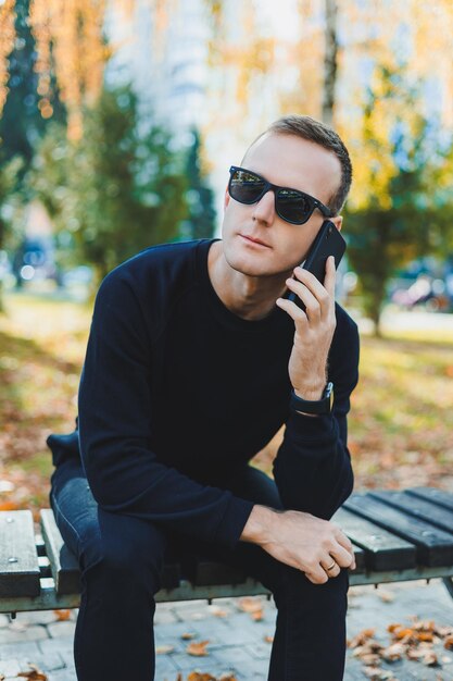 A lonely young man in a black sweater sits in the park on a\
bench and talks on the phone a warm autumn day