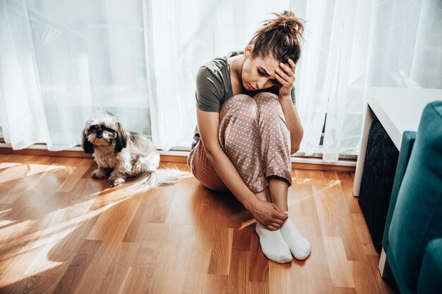 Lonely young woman in depression sit by window with embrace knees lost in bad pessimistic thoughts.