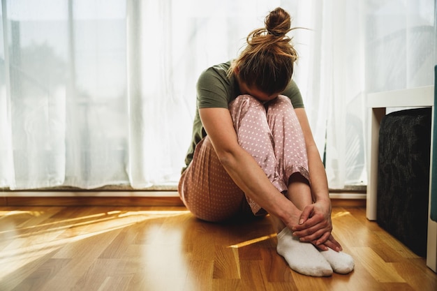 Lonely young woman in depression sit by window with embrace knees lost in sad thoughts.