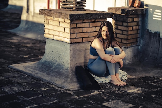 Lonely young woman in depression sit with embrace knees on a rooftop terrace lost in bad pessimistic thoughts after yoga training.