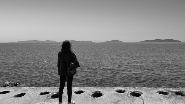 海岸で一人で立っており、地平線の白黒写真を見ている孤独な若い女性