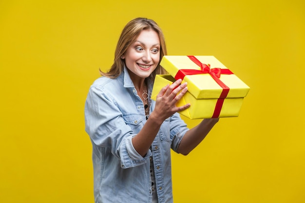 Долгожданный подарок. Портрет любопытной женщины в джинсовой рубашке, заглядывающей внутрь подарочной коробки, проверяющей, что внутри, распаковывающей с довольным счастливым выражением лица. студийный снимок на желтом фоне