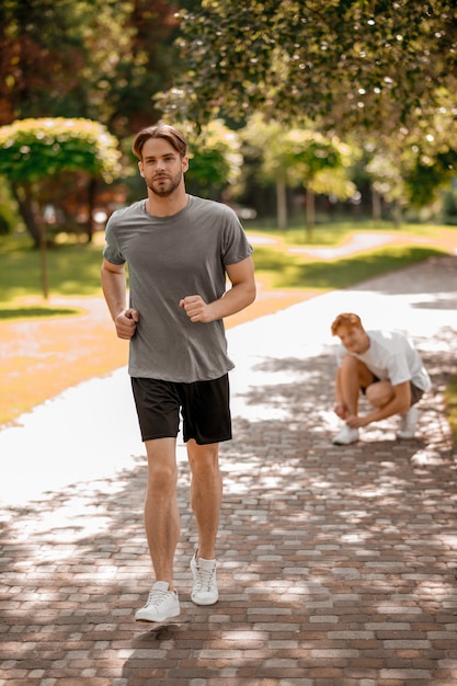 Corsa sulla lunga distanza. esecuzione di maratona di ragazzo barbuto dai capelli scuri e amico di zenzero sparpagliato accovacciato che si allaccia i lacci delle scarpe nel parco la mattina di sole