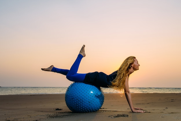 Long-haired blonde is engaged in Pilates on a training ball on the beach during sunset. Fit woman stretching her body using fitness ball. Concept of Health.