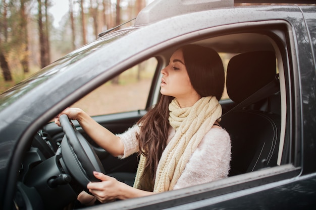 Long-haired brunette on the auto. A female model is wearing a sweater and a scarf. Autumn concept. Autumn forest journey by car.