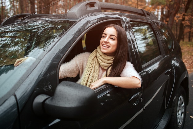 自動車の長い髪のブルネット。女性モデルがセーターとスカーフを着ています。秋のコンセプトです。車で秋の森の旅