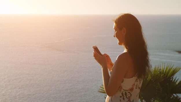 Bruna dai capelli lunghi si erge sul bordo infinito della costa dell'oceano blu e digita sullo smartphone moderno sotto la luce solare intensa