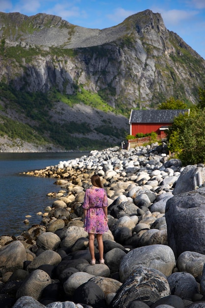 long-haired girl in purple dress stands on rocks with red house and massive mountains in background