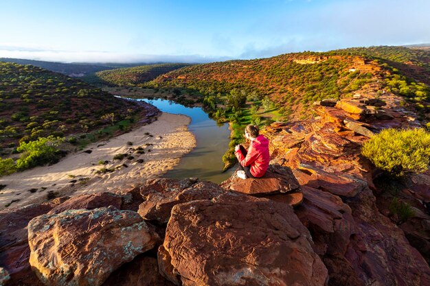 long-haired girl sits on top of hill on the red rocks of kalbarri national park in western australia