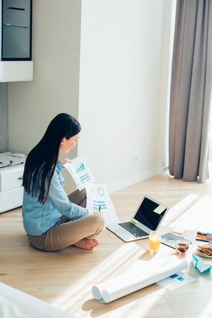 Long haired young lady sitting in front of a laptop with food on the floor and holding graphic charts