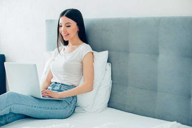 Giovane donna dai capelli lunghi seduta sul letto con un laptop moderno e guardando lo schermo banner del sito web