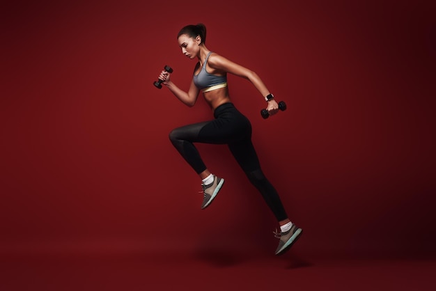 Long jump sportswoman jumping over red background with dumbbells in her hands