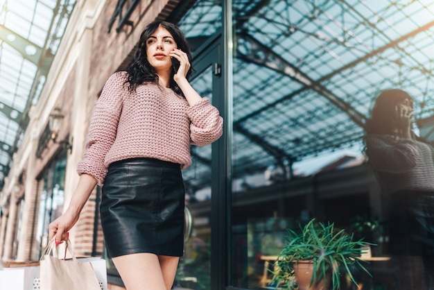 Long legged brunette woman in leather short skirt standing in front of shop window, holding a bunch of paper bags in hand and talking on mobile phone