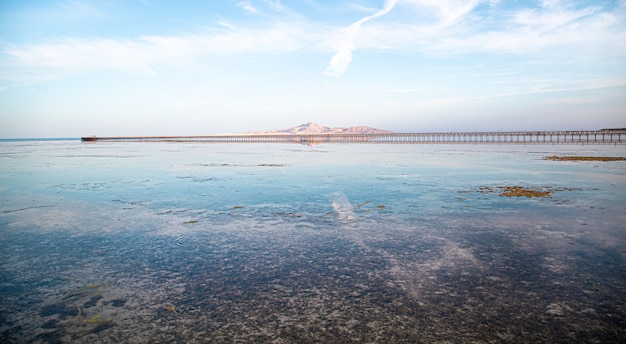 Длинный пирс среди моря и гор. Небо отражается в воде.