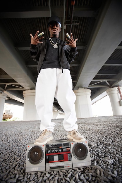 Long shot of cool rapper in trendy attire standing on top of old tape recorder in urban environment under bridge during performance