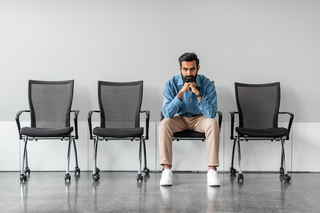 Foto lungo tempo di attesa pensieroso serio uomo d'affari indiano tenendo le mani sul mento seduto sulla sedia