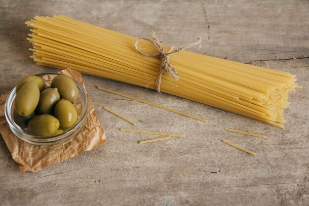 Long yellow raw spaghetti and green olives in glass bowl on wooden background