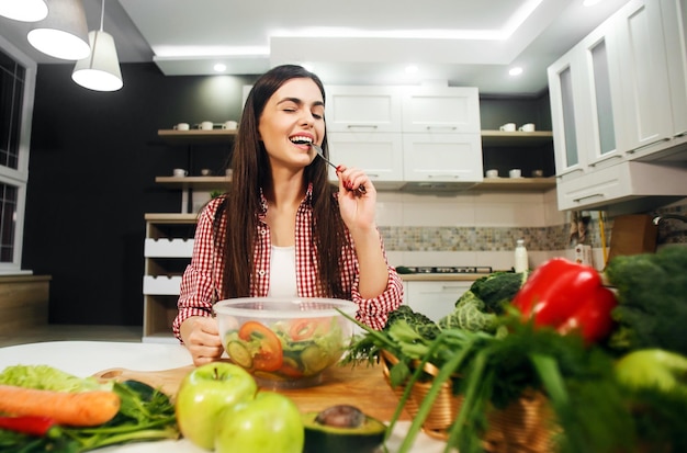 Longhaired caucasian girl enjoying healthy mixed salad wearing cool checked red shirt indoor shot in light big kitchen with table full of vegetables