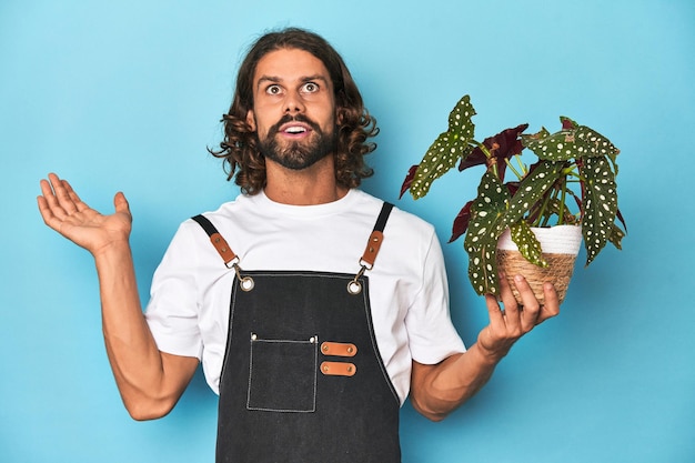 Longhaired gardener with beard holding a plant surprised and shocked