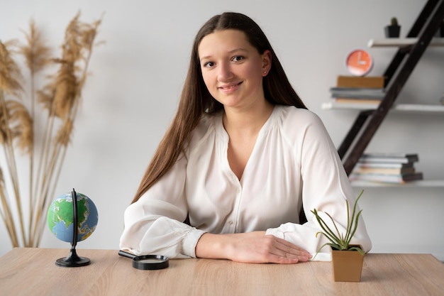 A longhaired girl student sitting at a desk in the office listens to or watches an online lesson while studying remotely A college student is looking at the camera Learning and education concept