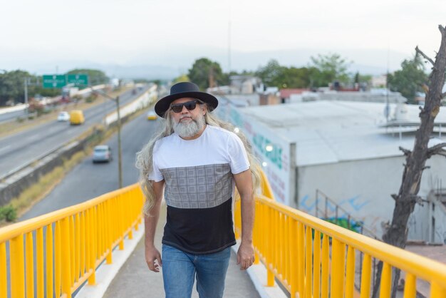 Longhaired grayhaired man wearing a black hat on a pedestrian bridge overlooking the city