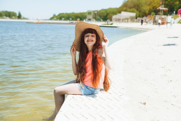 Длинноволосая маленькая девочка в пляжной шляпе сидит на пляже и смотрит на море