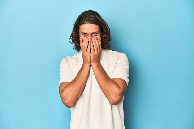 Foto uomo dai capelli lunghi in uno studio blu polo bianco che ride di qualcosa che copre la bocca con le mani