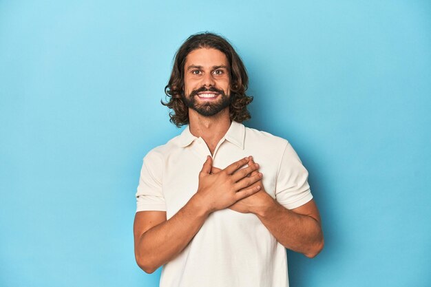 Photo longhaired man in a white polo blue studio laughing keeping hands on heart concept of happiness