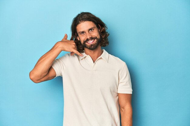 Photo longhaired man in a white polo blue studio showing a mobile phone call gesture with fingers