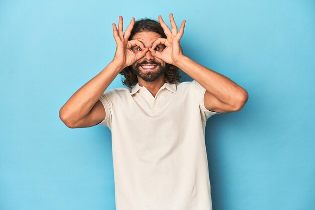 Photo longhaired man in a white polo blue studio showing okay sign over eyes